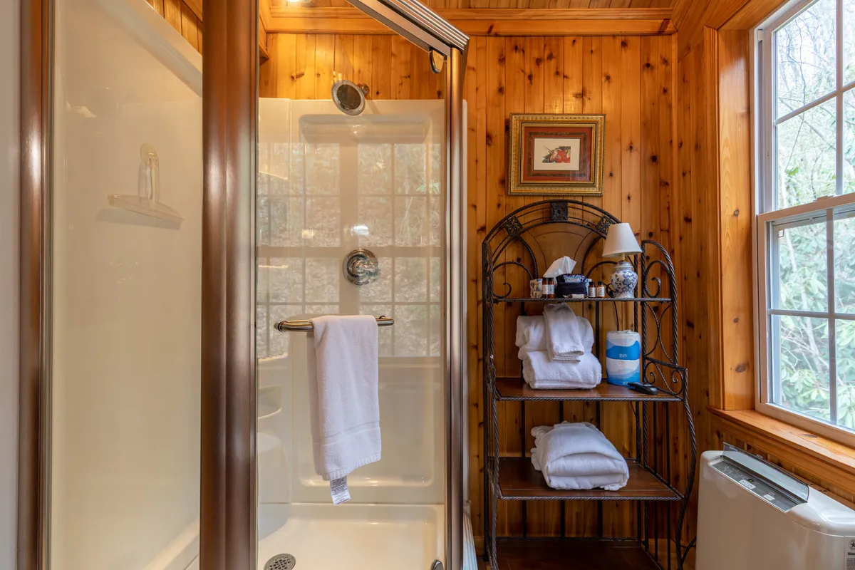 A modern, serene bathroom featuring a glass shower enclosure, a towel rack, a window letting in natural light, and a piece of decorative art. The space feels fresh, clean, and inviting—ideal for self-care routines and refreshing after a day of adventure.