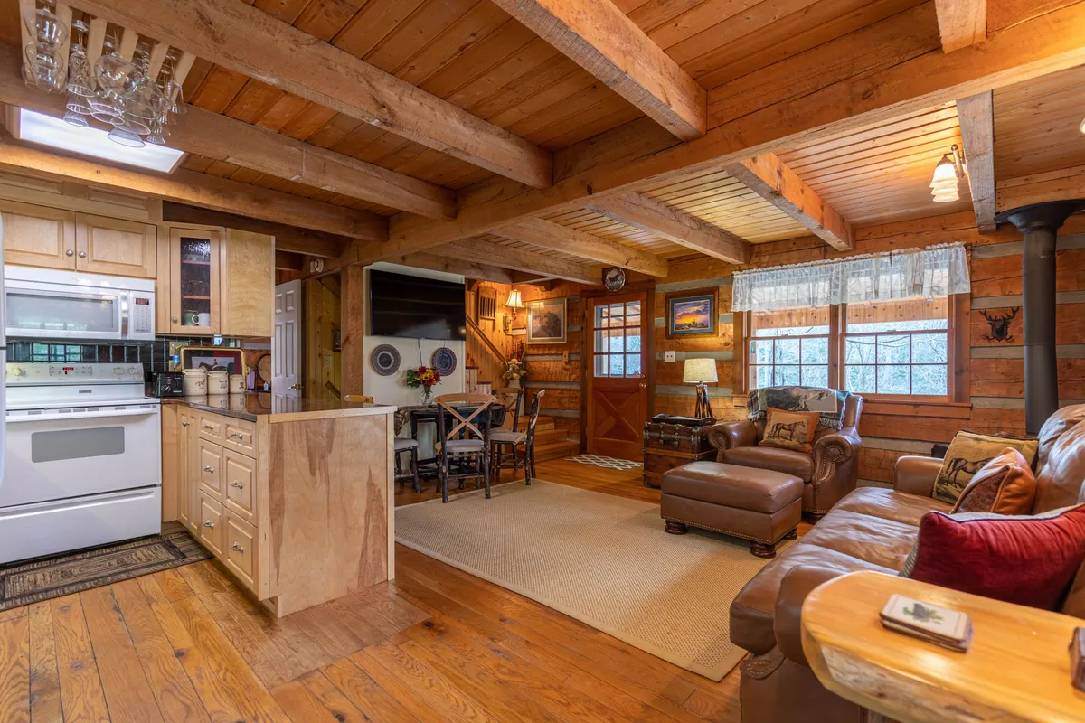 This photo captures the open living area with rich wooden beams, a comfortable leather sofa, a dining area, and a glimpse of the kitchen counter. The warmth of the space and the inviting seating make it ideal for gatherings, family movie nights, or relaxing by the fire.