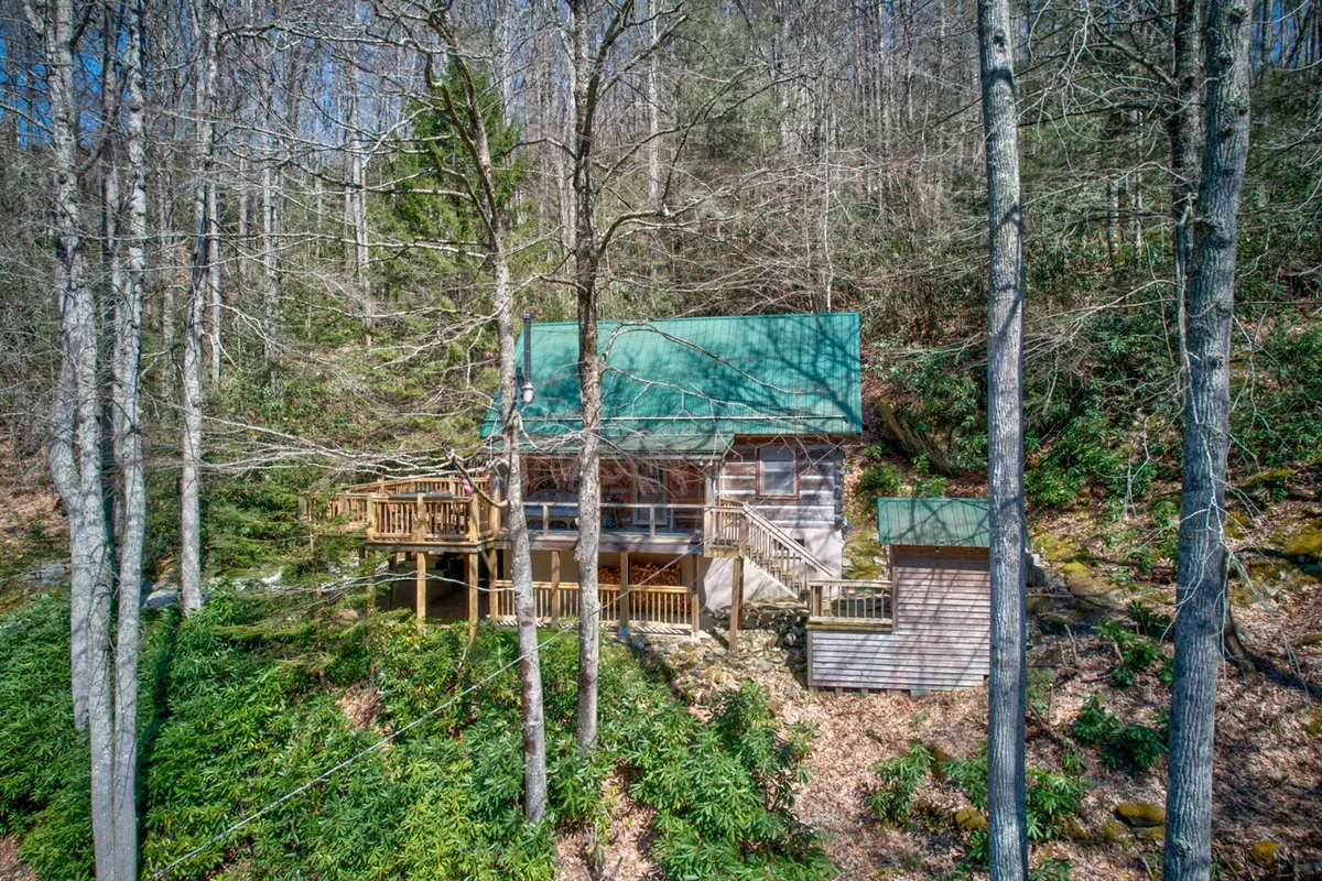The image features the Creeksong Cabin nestled among dense trees, with its green metal roof and spacious wooden deck standing out. The setting is serene and surrounded by nature, making the cabin appear secluded and inviting. The vantage point suggests easy access to outdoor activities like nature walks, wildlife watching, and relaxing on the deck.