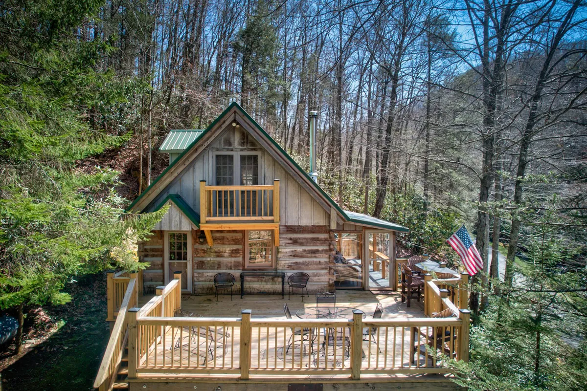 A broad view of the wooden deck attached to an A-frame cabin, accented by an American flag. The deck provides multiple seating options for relaxing, with views of the forested landscape and the suggestion of an upper balcony for even more private moments. The atmosphere is both cozy and welcoming, suggesting leisurely mornings or evenings spent outdoors.