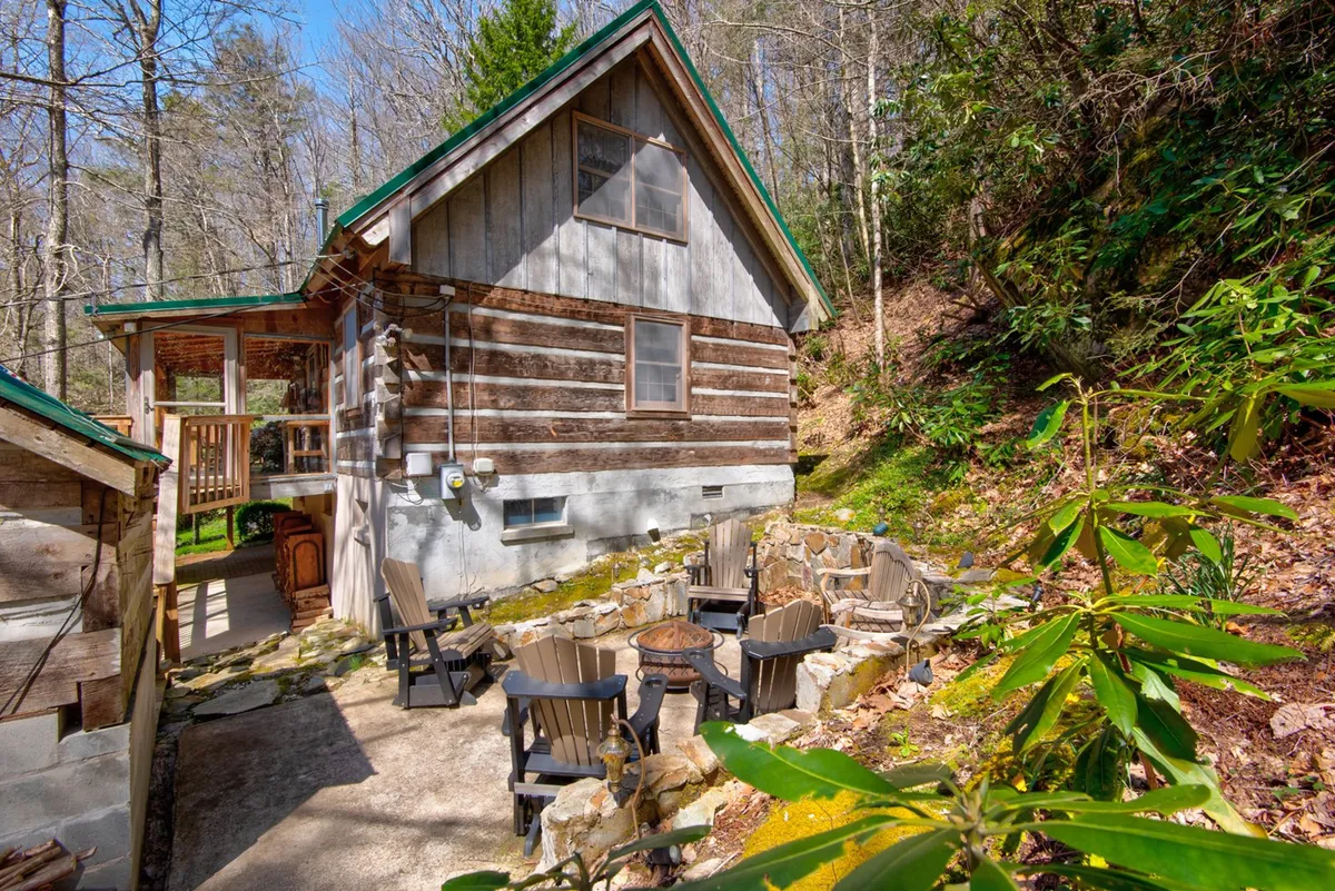 This image showcases the log cabin’s exterior nestled in lush natural surroundings, featuring a spacious seating area with a fire pit. The arrangement encourages outdoor gatherings, storytelling, and marshmallow roasting as the sun sets behind the trees.