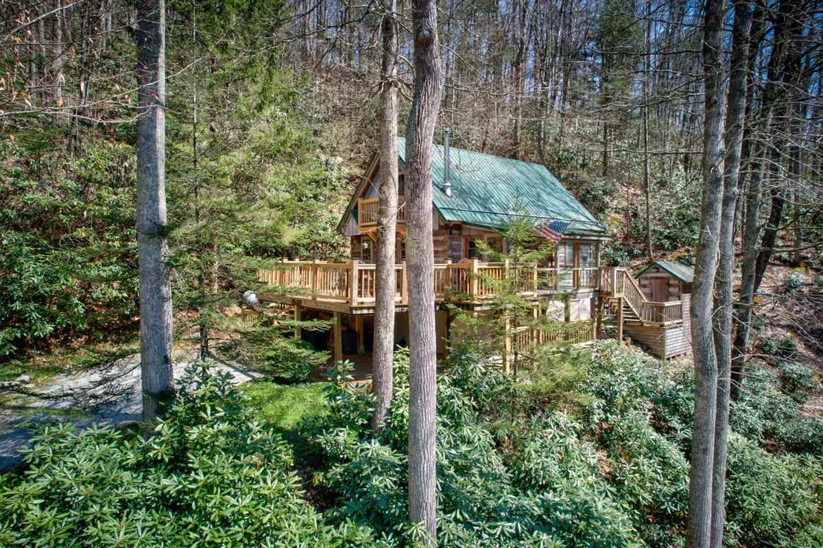 The wooden cabin is nestled among tall trees, with a spacious deck area extending into the peaceful wilderness. The structure is framed by lush greenery, illustrating both seclusion and the inviting outdoor living space where guests can unwind and take in the scenic beauty.