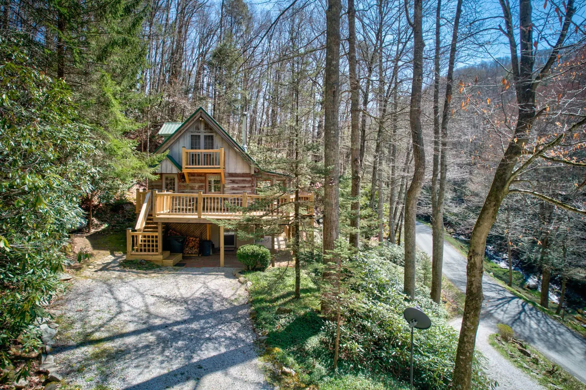 The image showcases a charming wooden cabin nestled among lush trees, with a welcoming balcony overlooking a large gravel driveway. The cabin is surrounded by natural beauty, offering tranquil views of the forest and hints of the nearby creek. Soft golden light at sunset filters through the trees, creating a warm and inviting atmosphere perfect for relaxation and outdoor enjoyment.