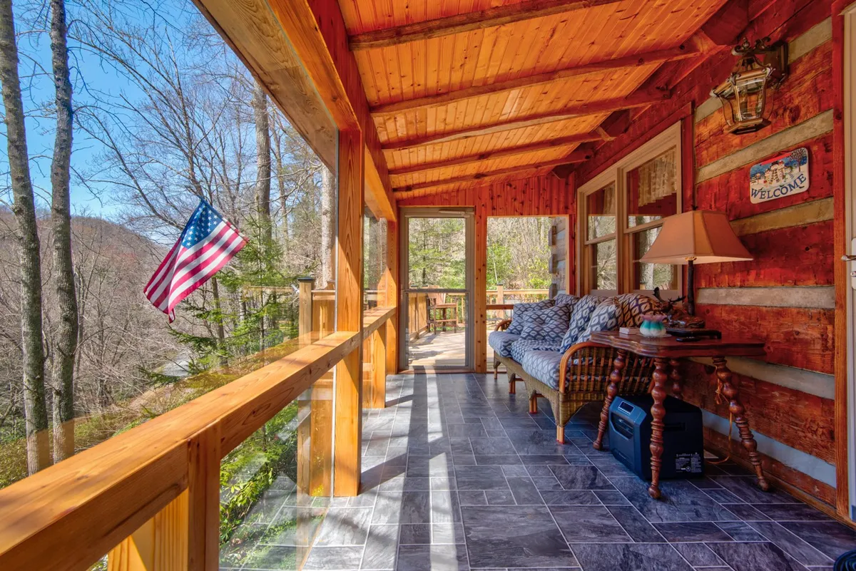 This interior scene features a cozy sofa with patterned cushions, a wooden table, and an American flag accent. Large windows allow sunlight to filter in, and a lantern-style light fixture adds charm. The comfortable setup is ideal for reading or relaxing while enjoying views of the outdoors, reflecting the inviting warmth of a sunroom.