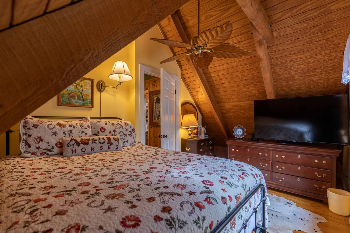 A cozy bedroom featuring a queen bed with a floral quilt, exposed wooden beams overhead, a flat-screen television, and a classic wooden dresser adorned with decorative items. Soft natural light filters in through the window, creating a tranquil and homey atmosphere.