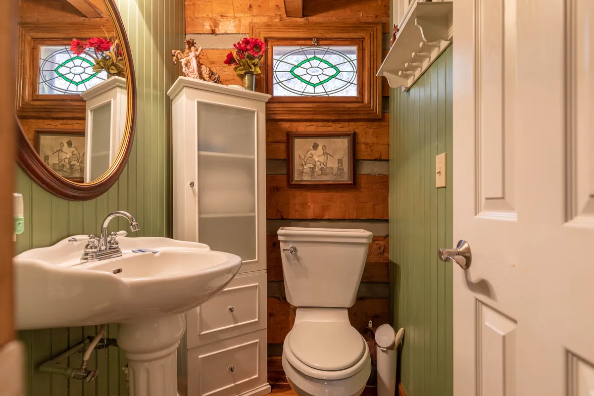 A welcoming half-bathroom with a round mirror, white sink, toilet, glass cabinet, and a stained glass window adding a pop of color. Fresh flowers and nature-inspired details create a warm, inviting mood.