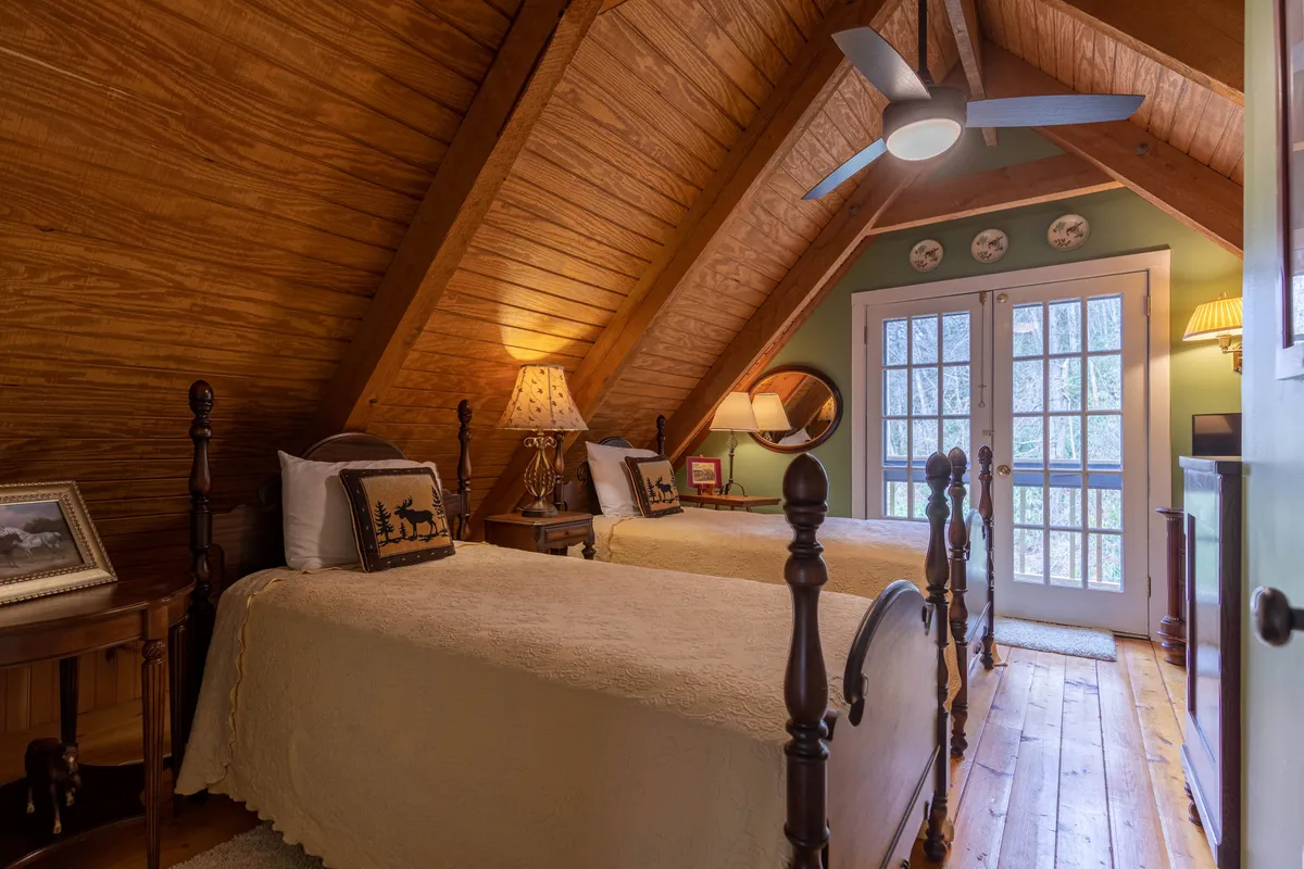 A charming bedroom with two twin beds positioned under a warm wooden ceiling, flanked by French doors that likely open to a Juliette balcony. A decorative lamp and thoughtfully arranged art create a welcoming and intimate space, perfect for kids, friends, or siblings.